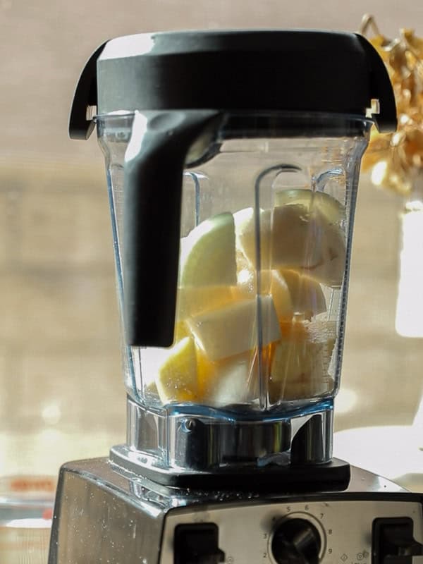 A blender filled with chopped apples, placed on a kitchen counter. Sunlight streams from a nearby window, casting shadows and highlighting the fresh fruit inside the transparent blender jar.